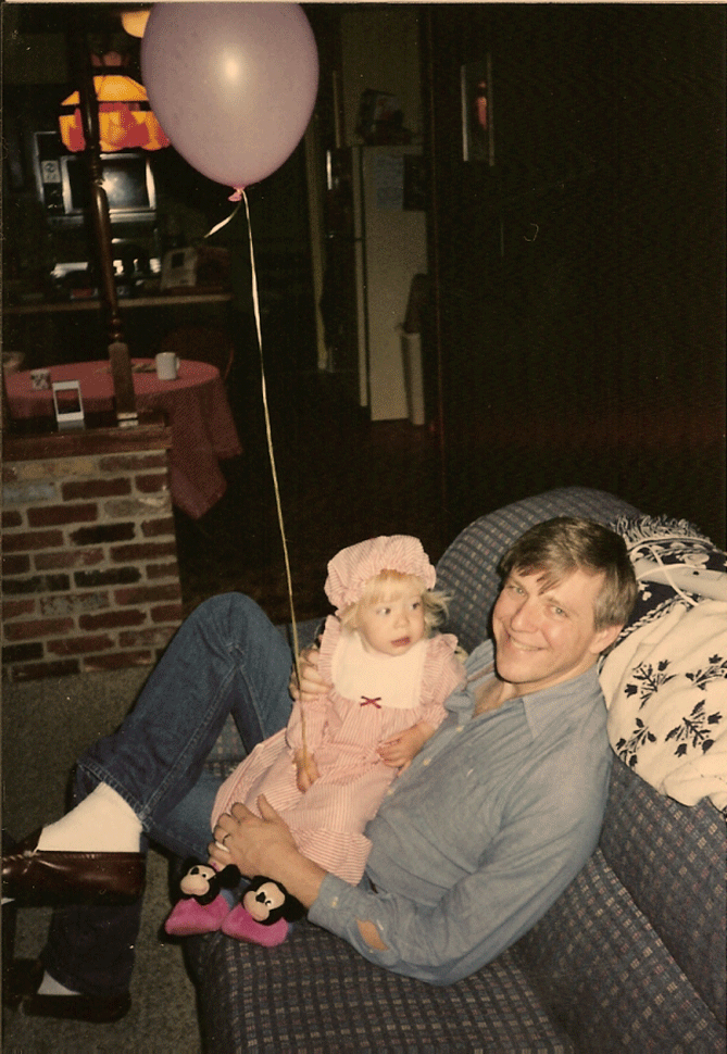 A very, very young Rachel, maybe three years old, is held by her dad on a couch. She has a balloon tied to her wrist, floating above and is wearing a matching bibbed dress and bonnet with mickey mouse slippers