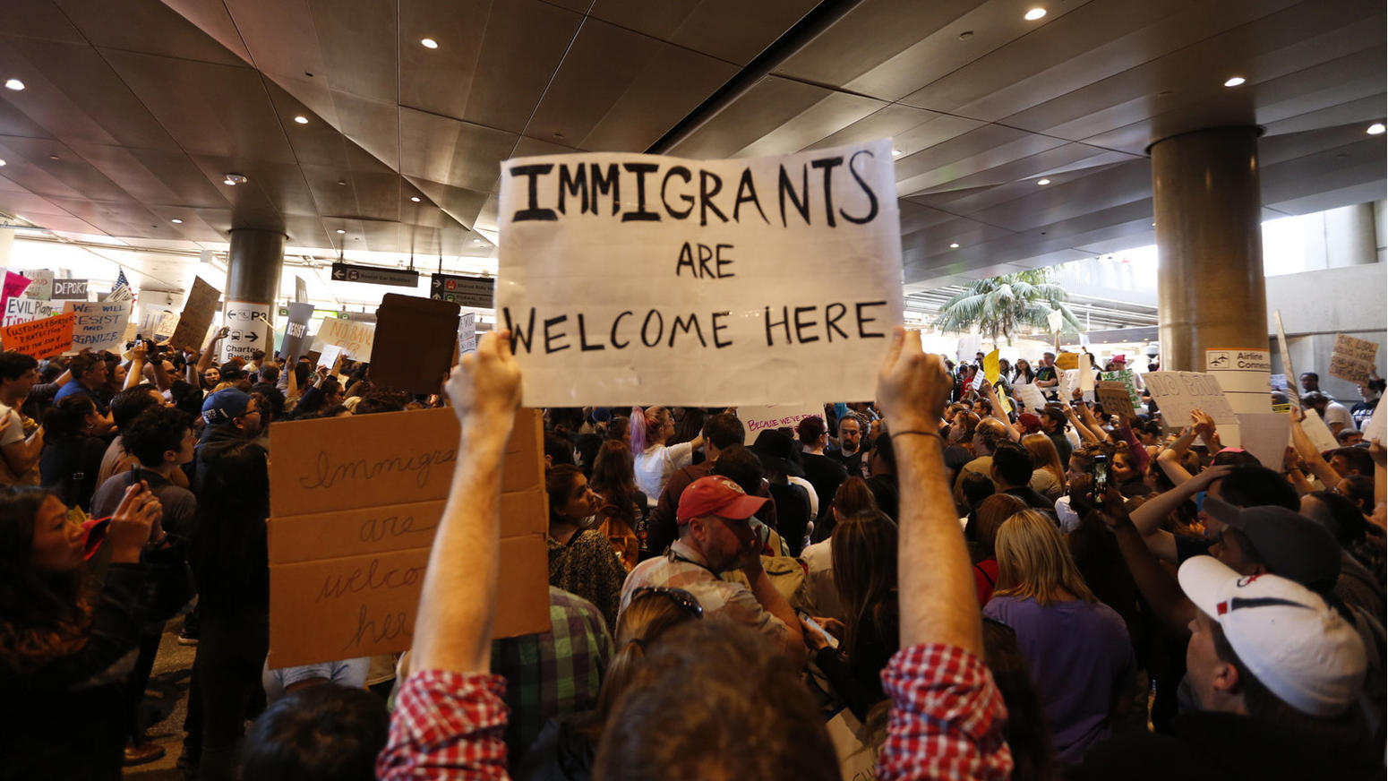 A woman holds up a black and white sign in a large crowd at an airport protest that reads 