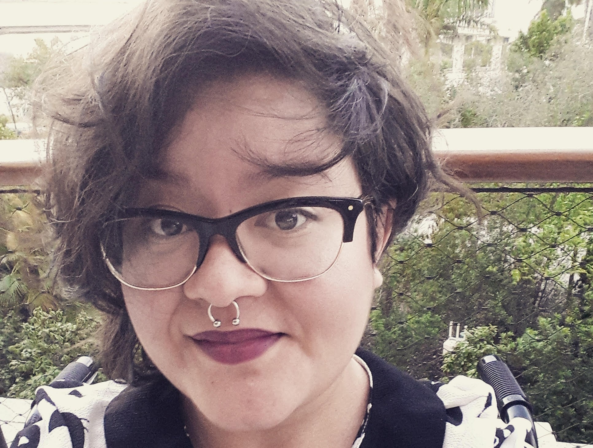 A Latinx person with short black hair wearing red lipstick, black framed glasses, and a pin that says 