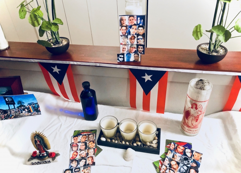 A home photograph of an altar built in a living room with a collection of white table cloth, candles, and Puerto Rican flags.