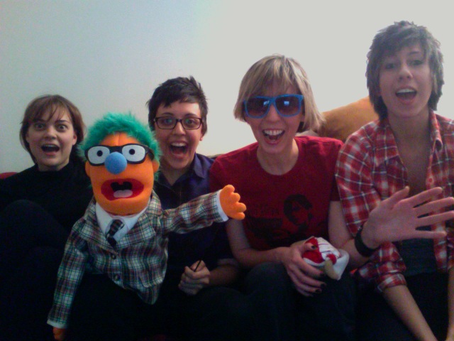 A throwback picture of the early days of Autostraddle. Robin Roemer (wearing a black turtle neck and brown hair with bangs), Carly Usdin (wearing a blue button-up, short brown hair, and glasses), Riese Bernard (wearing a red t-shirt, blue sunglasses, and a blonde gay haircut), and Alex Vega (wearing a red plaid shirt and a brown very gay haircut) sit on a couch, all with their mouths excitedly agape. Carly is holding a bright orange puppet guy with glasses, teal hair, and a blue nose.