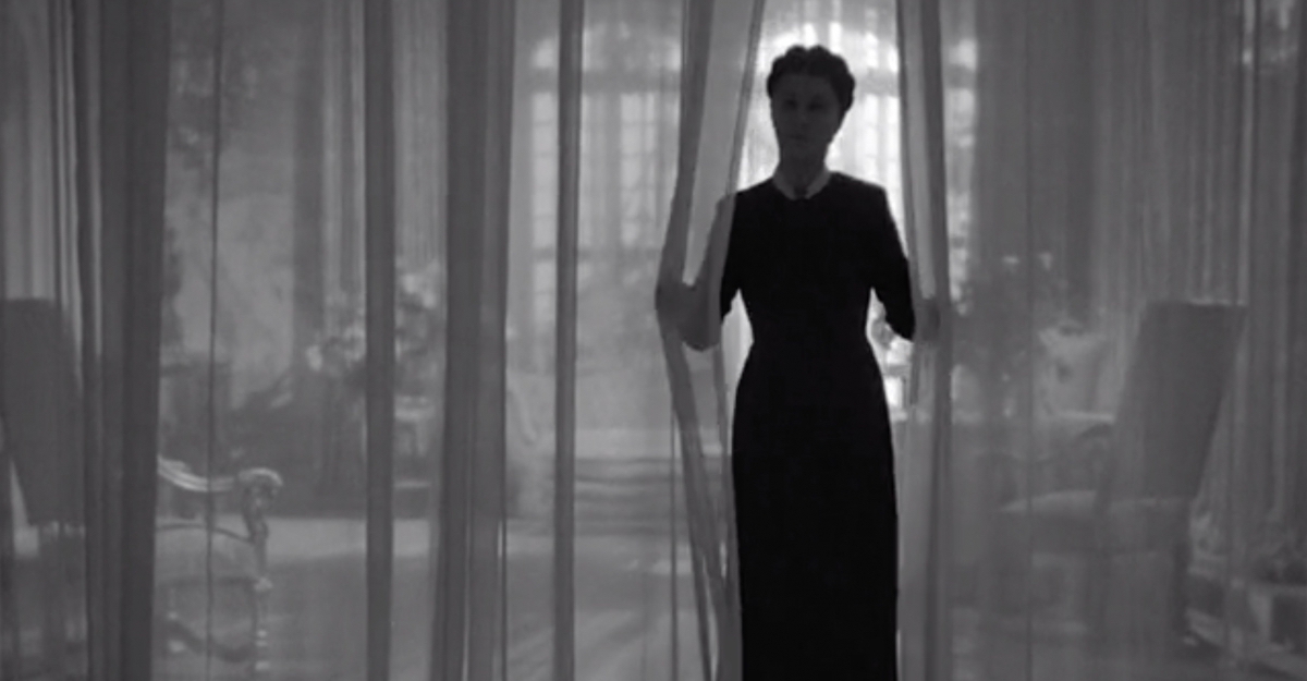 The shadowy figure of Mrs. Danvers appearing through a pair of curtains.