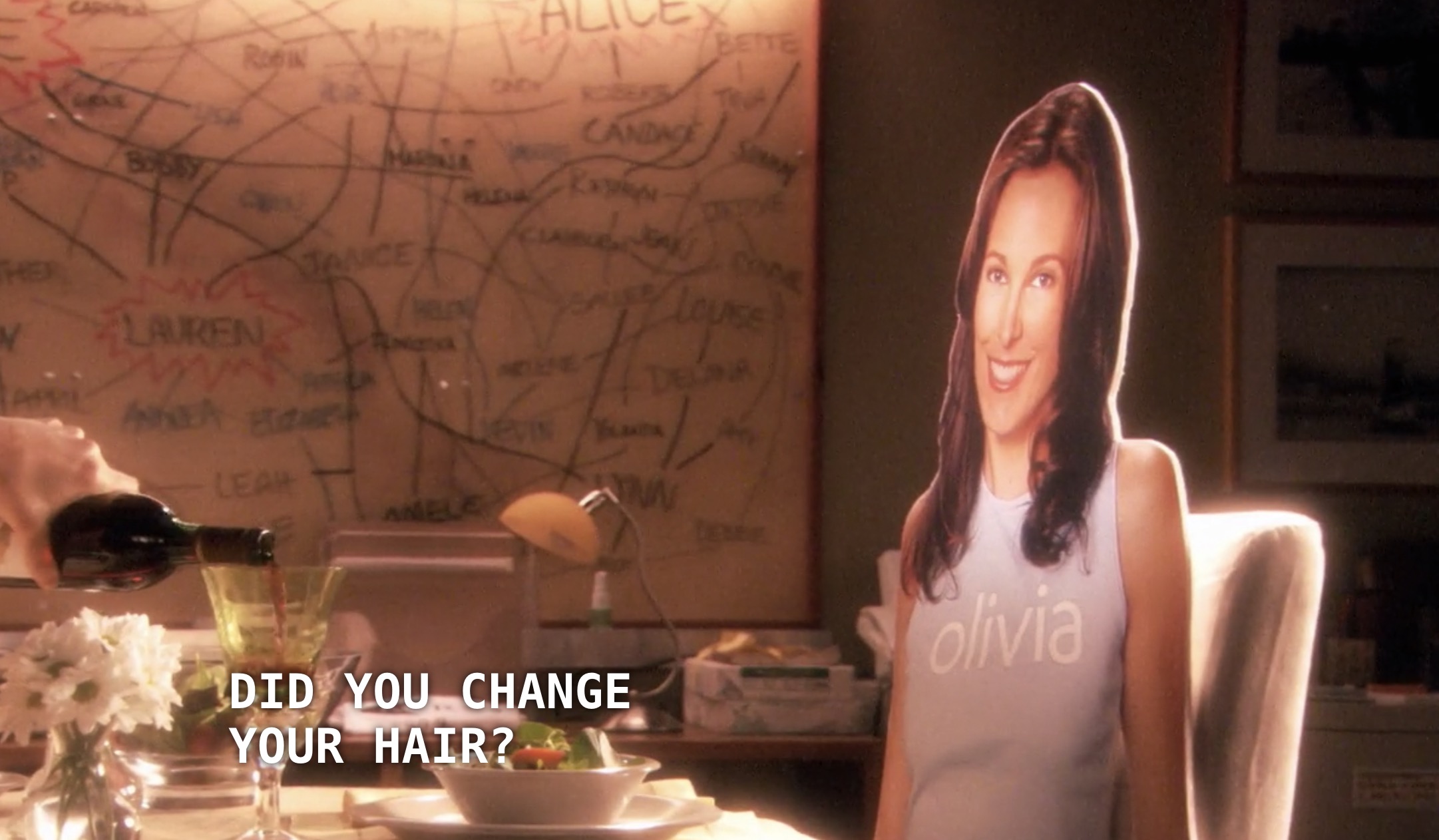 A cardboard cutout of Dana (wearing a purple "Olivia" shirt) sits at a table that is set with wine, flowers, and salad. Alice's massive Our Chart is hanging on the wall in the background. Alice is off screen except for her hand, which is pouring a glass of wine. Alice says, "Did you change your hair?"