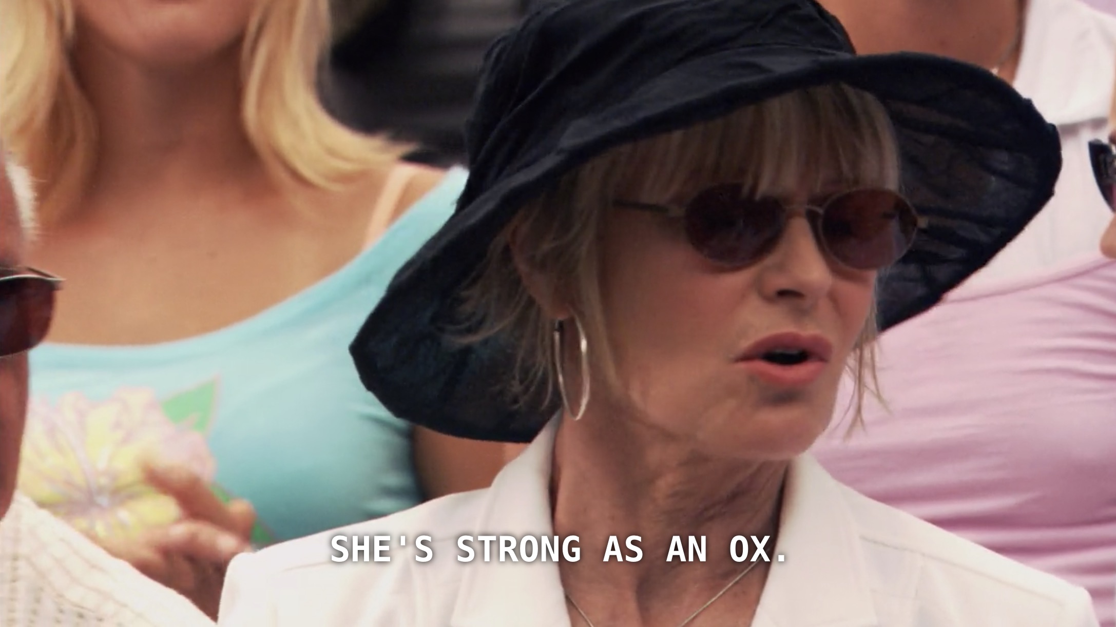 Dana's mom (wearing a cream blazer and a black floppy hat) sits in the stands of Dana's tennis match. She says, "she's strong as an ox" in reference to her daughter's tennis skills.