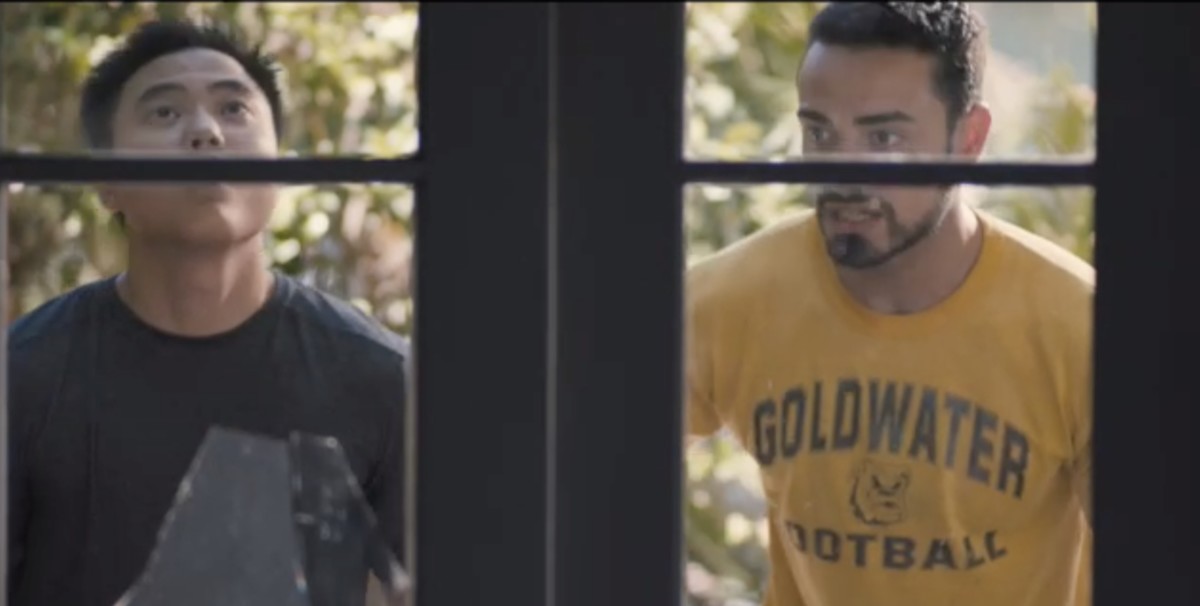 Leo and Jose stand in front of Jose's door. One window pane is broken. Jose is wearing a yellow t-shirt.