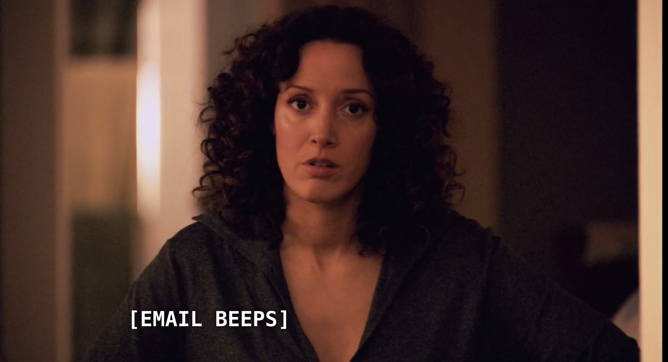 Bette (with curly dark hair and a black deep-v blouse) is in her house. She just finished meditating. She hears a beep come from her email.