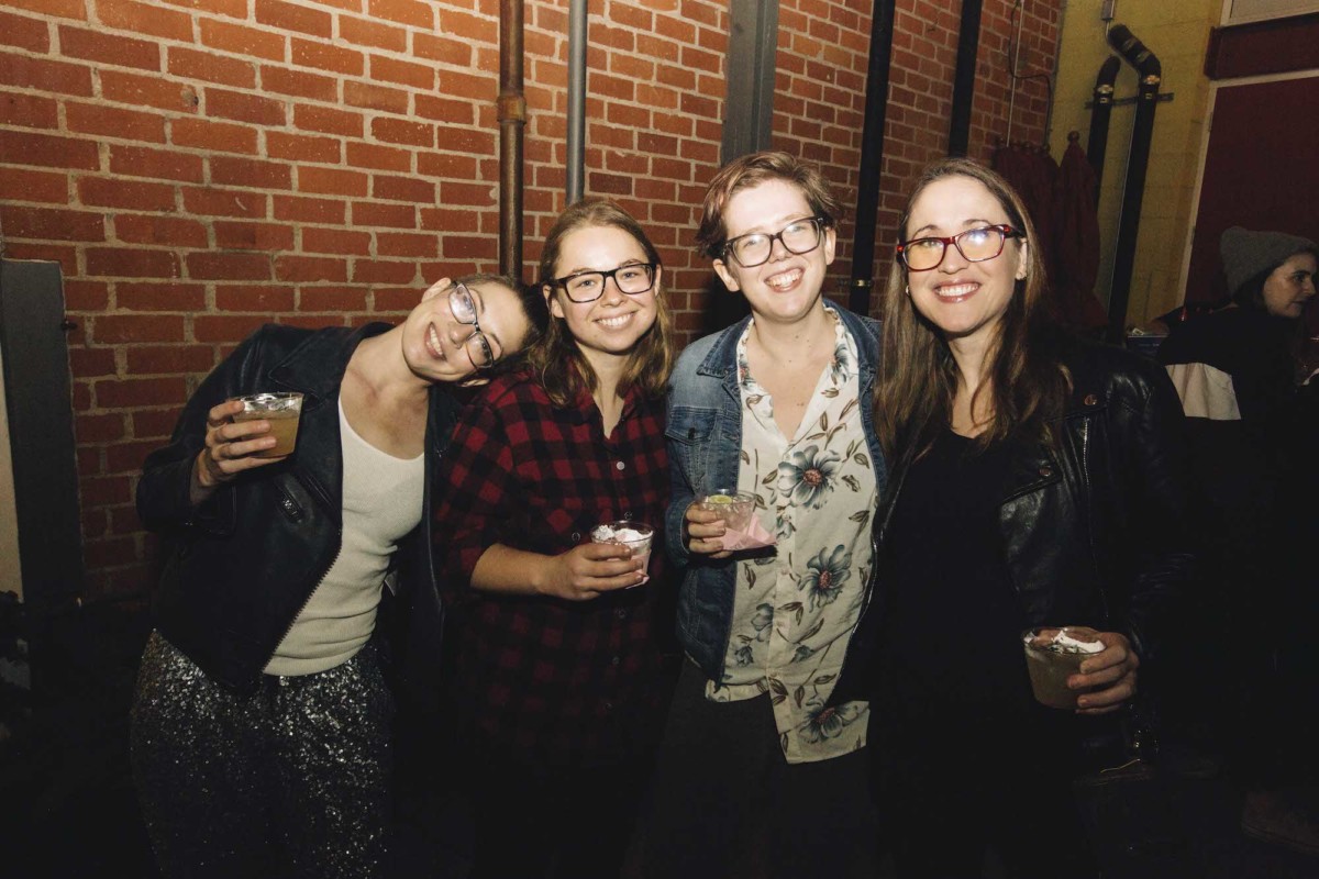 Four people holding drinks pose for a photo against a brick wall inside.