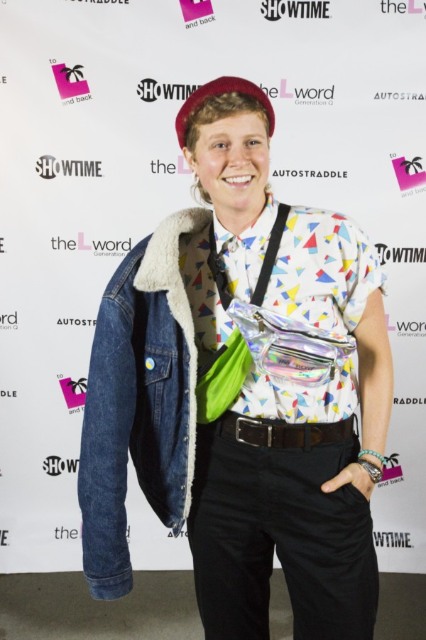 A person posing in front of a promo poster for "The L Word: Generation Q." They are wearing a brightly colored patterned button-up, two fanny packs crossing their chest, and a denim jacket draped over their right shoulder.