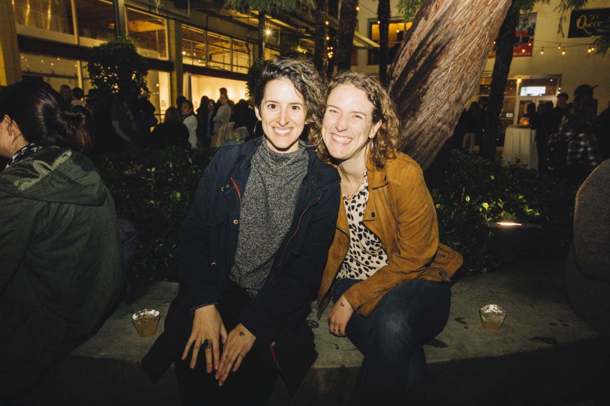 Two people sit outside on a bench and pose for a photo, smiling.