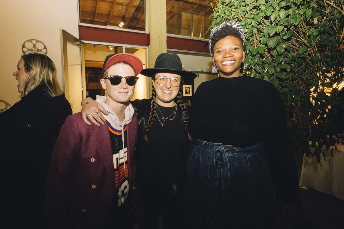Three people stand next to each other, posing for a photo. The one on the left is wearing a maroon jacket, sunglasses, and a baseball hat. The person in the middle is wearing all black, two braids in their brown hair, and glasses. The person on the right is wearing a high-waisted denim skirt, a black top, and a headscarf.
