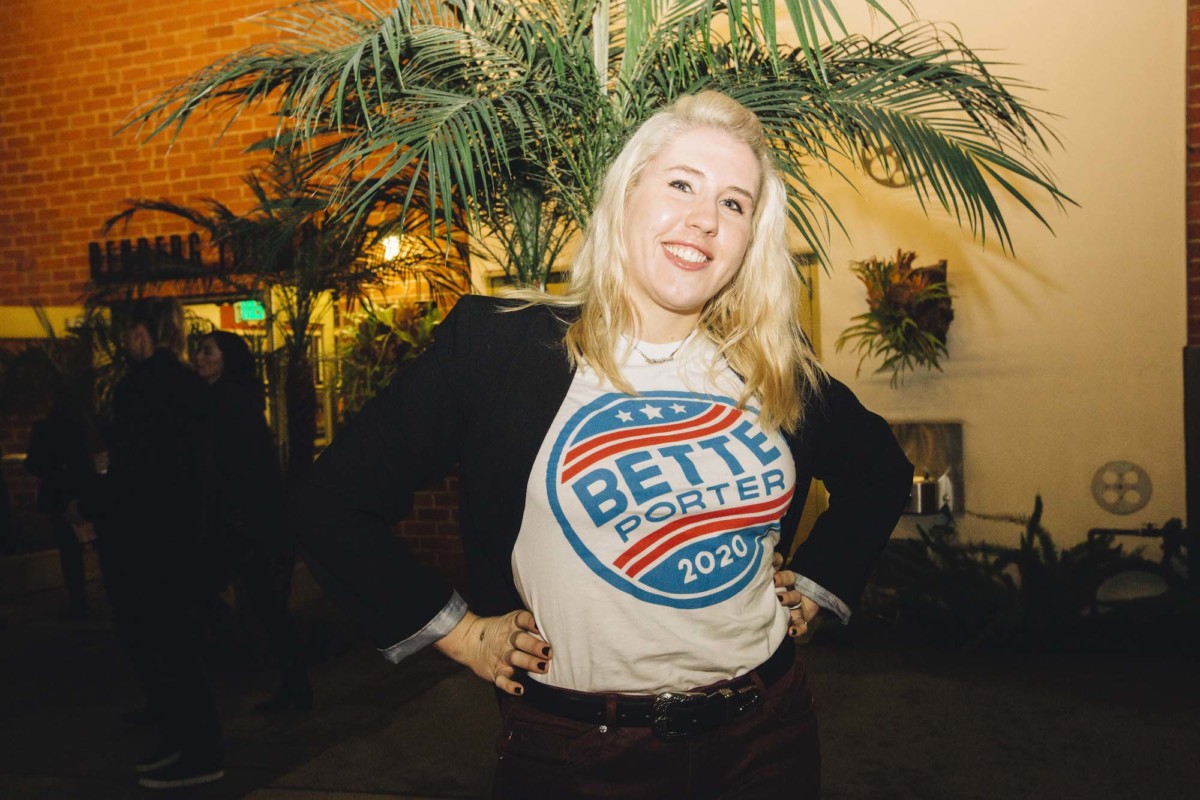 A blonde-haired person wearing a "Bette Porter 2020" t-shirt under a black blazer. They are smiling in front of a palm tree.