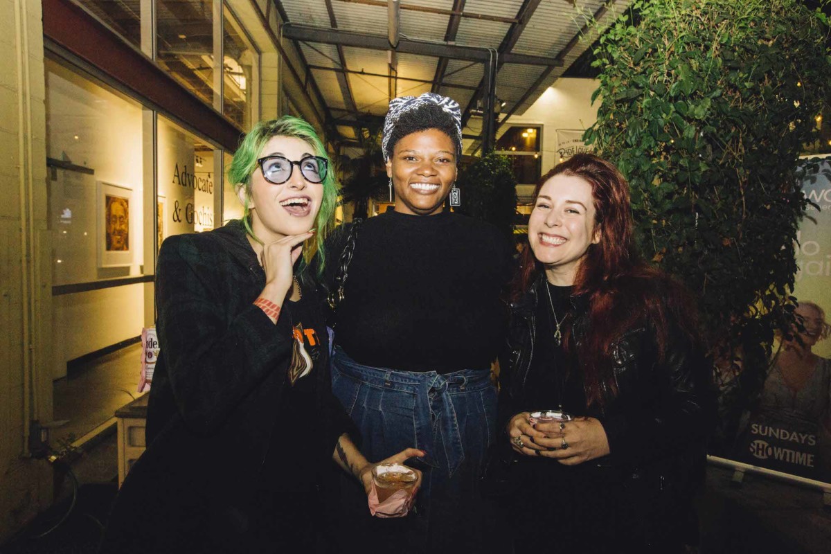 Three people posing for a photo. The one on the left has green hair and is wearing all black and glasses. The person in the middle is wearing a denim skirt, black top, and a headscarf. The person on the right has long dark red hair and is wearing all black.