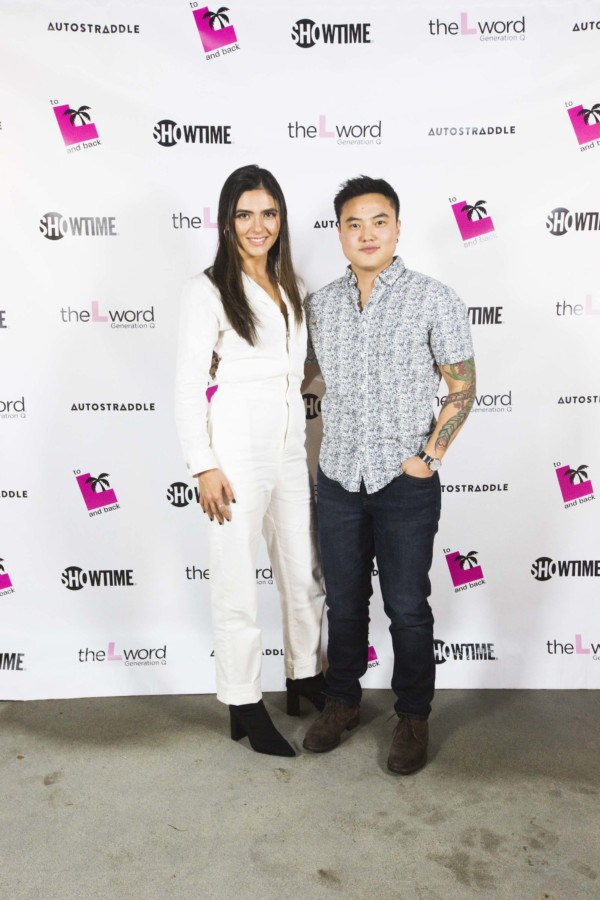 Arienne Mandi (in a white jumpsuit and black ankle boots) and Leo Sheng (in dark jeans and a black-and-white patterned button-up shirt) posing in front of a promo poster for "The L Word: Generation Q."