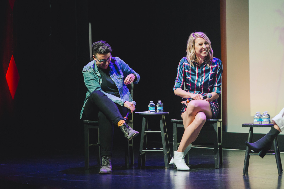 The "To L and Back" hosts Carly Usdin and Riese Bernard sit next to each other on stage. Carly is wearing black jeans and a chambray shirt, and Riese is wearing a blue, red, and white striped dress.