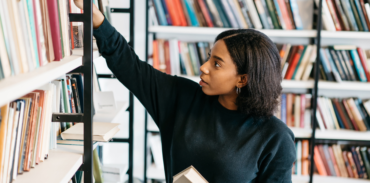 woman in a library holding a book and reaching for another on a shelf