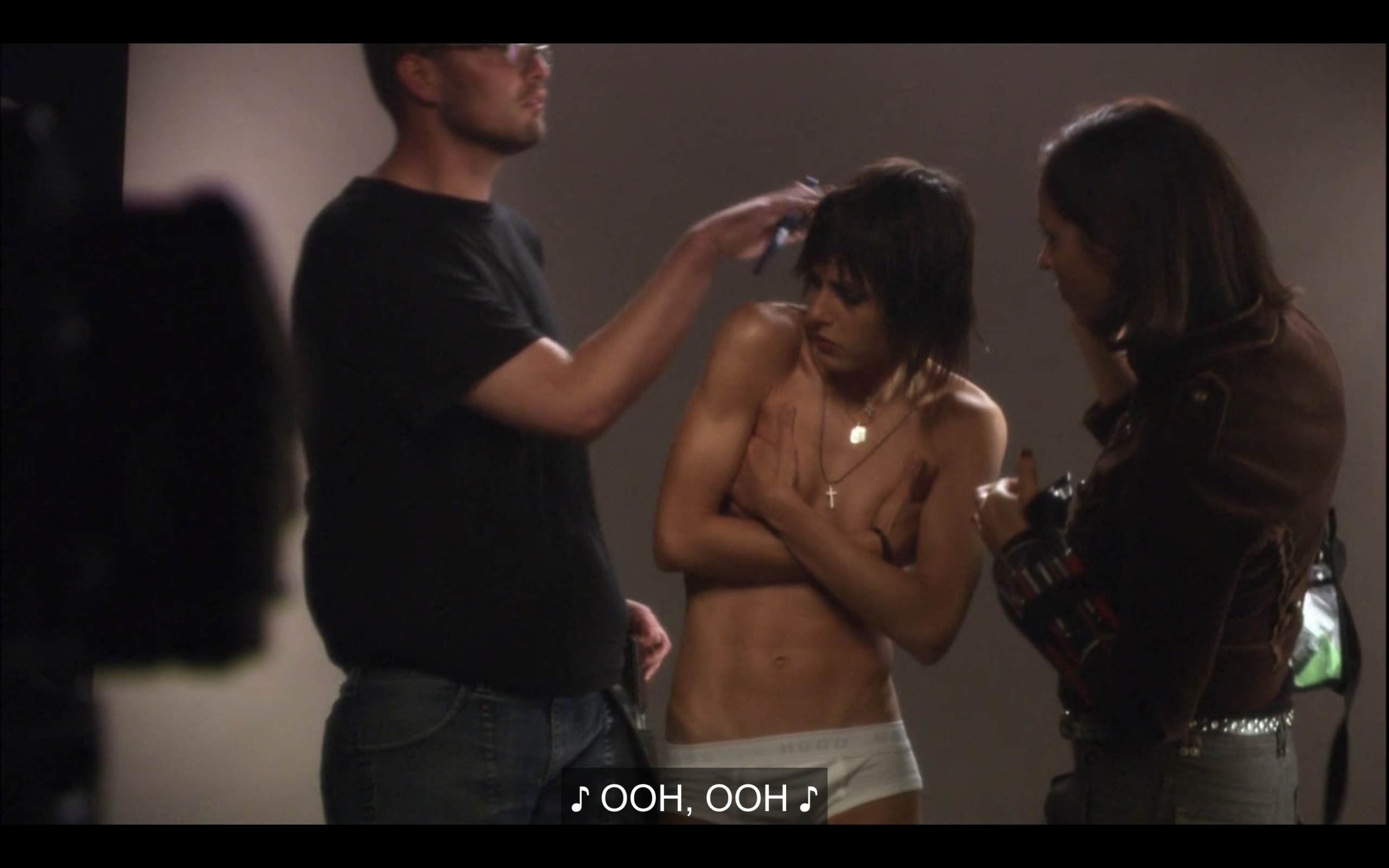 Shane stands in white brief underwear and topless for a photoshoot. Her hands are covering her boobs, as two people prep her for a photoshoot. 