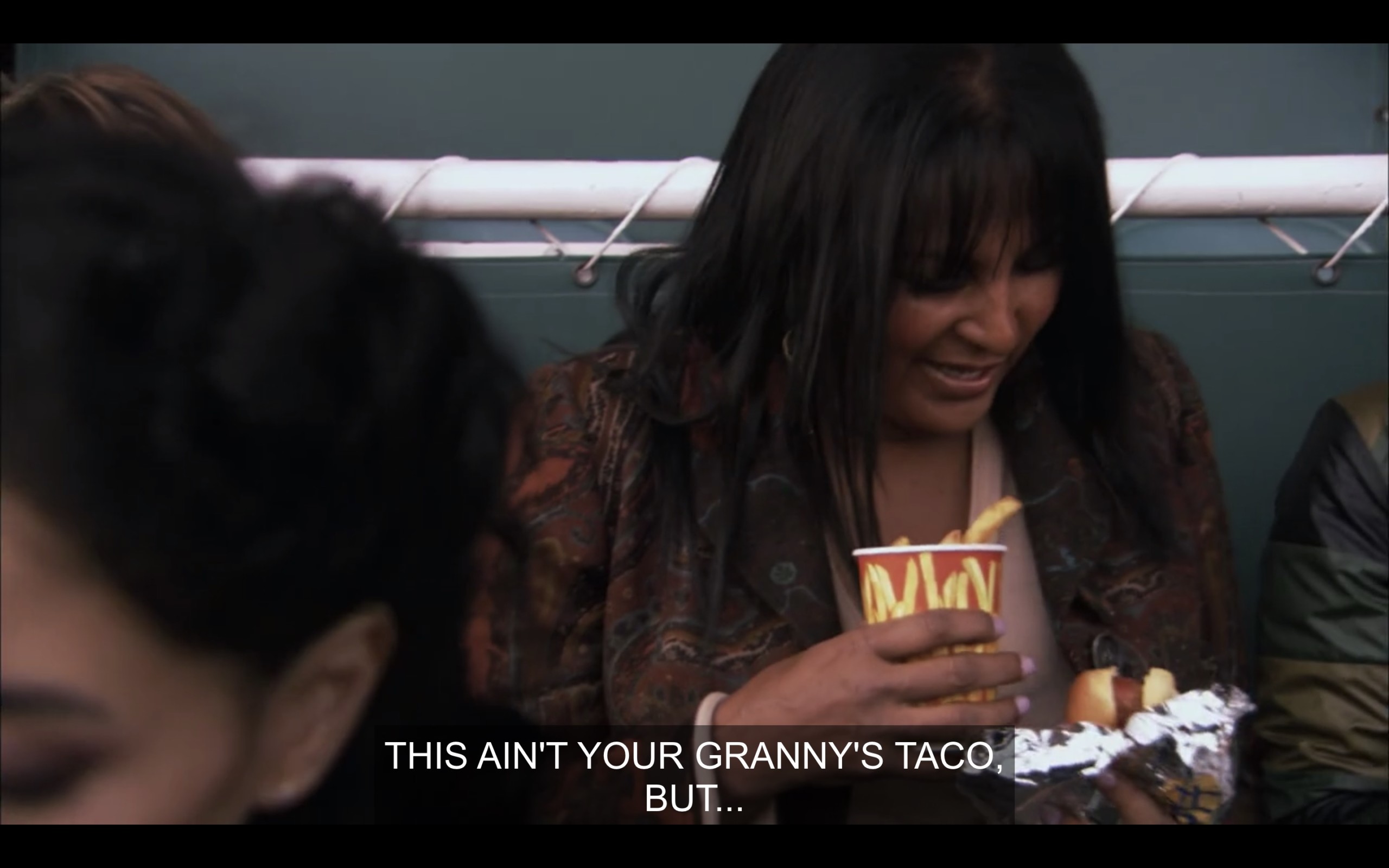 Kit holding a cup of fries in one hand and a hot dog wrapped in foil in another. She says, "This ain't your granny's taco, but..."