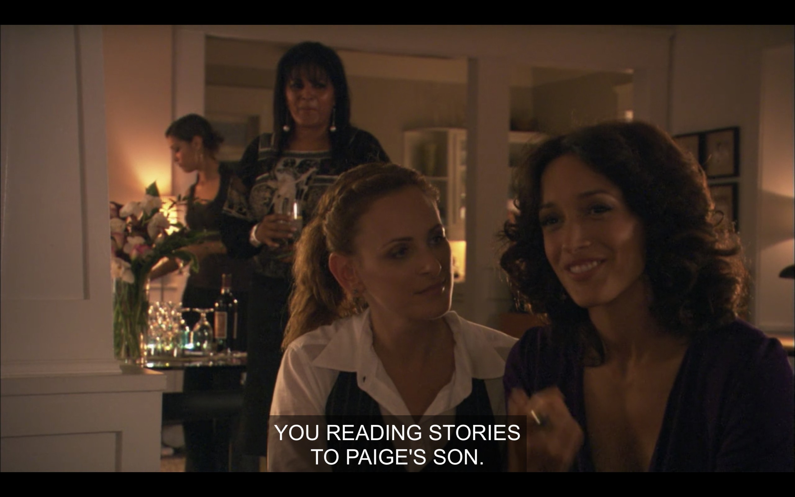 Jodi and Bette sitting on the couch, with Kit and Jenny standing off in the background. Bette says, "You reading stories to Paige's son."