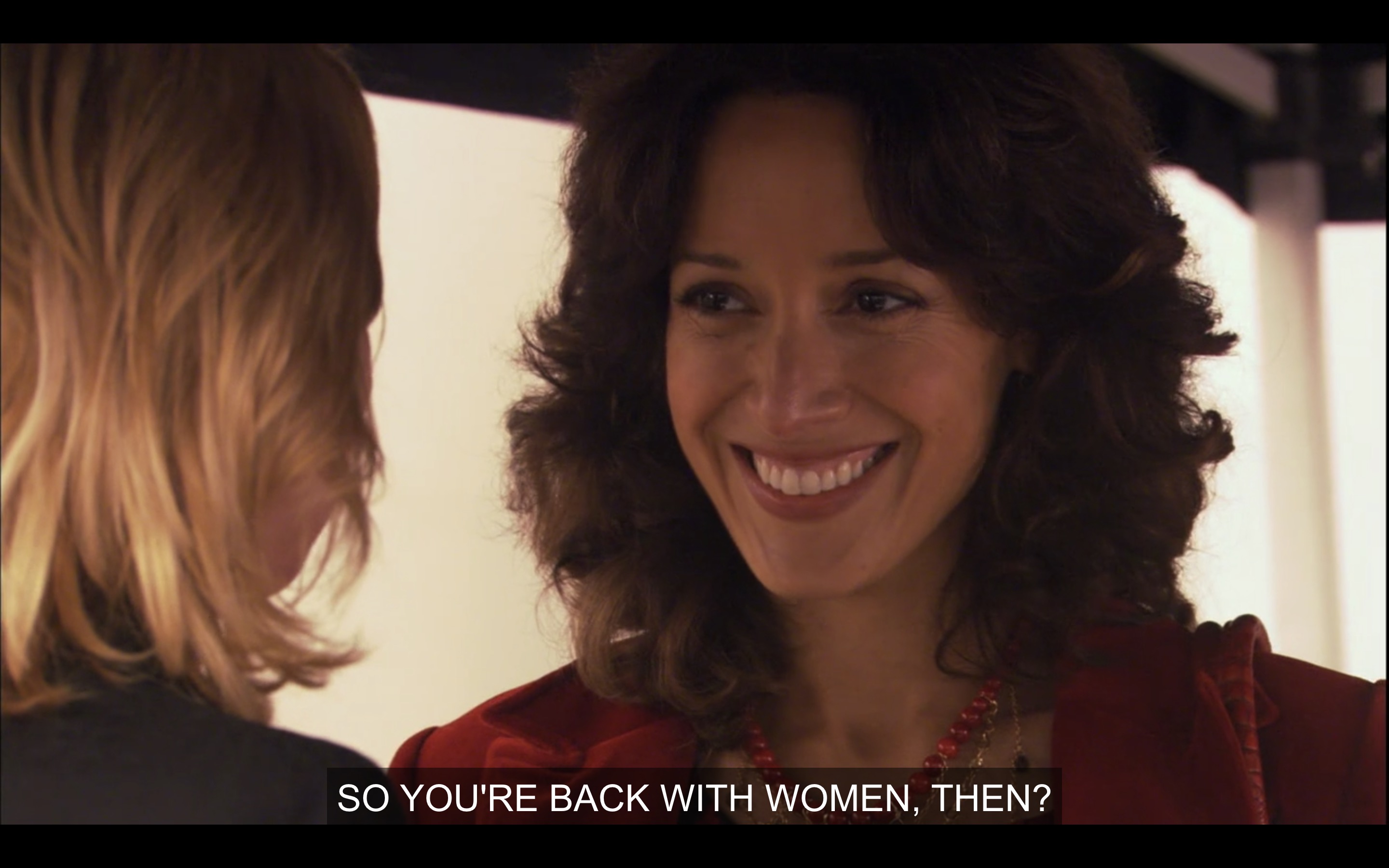 Bette (in a red velvet blazer) smiles big at Tina (whose back is to the camera). Bette says, "So you're back with women, then?"