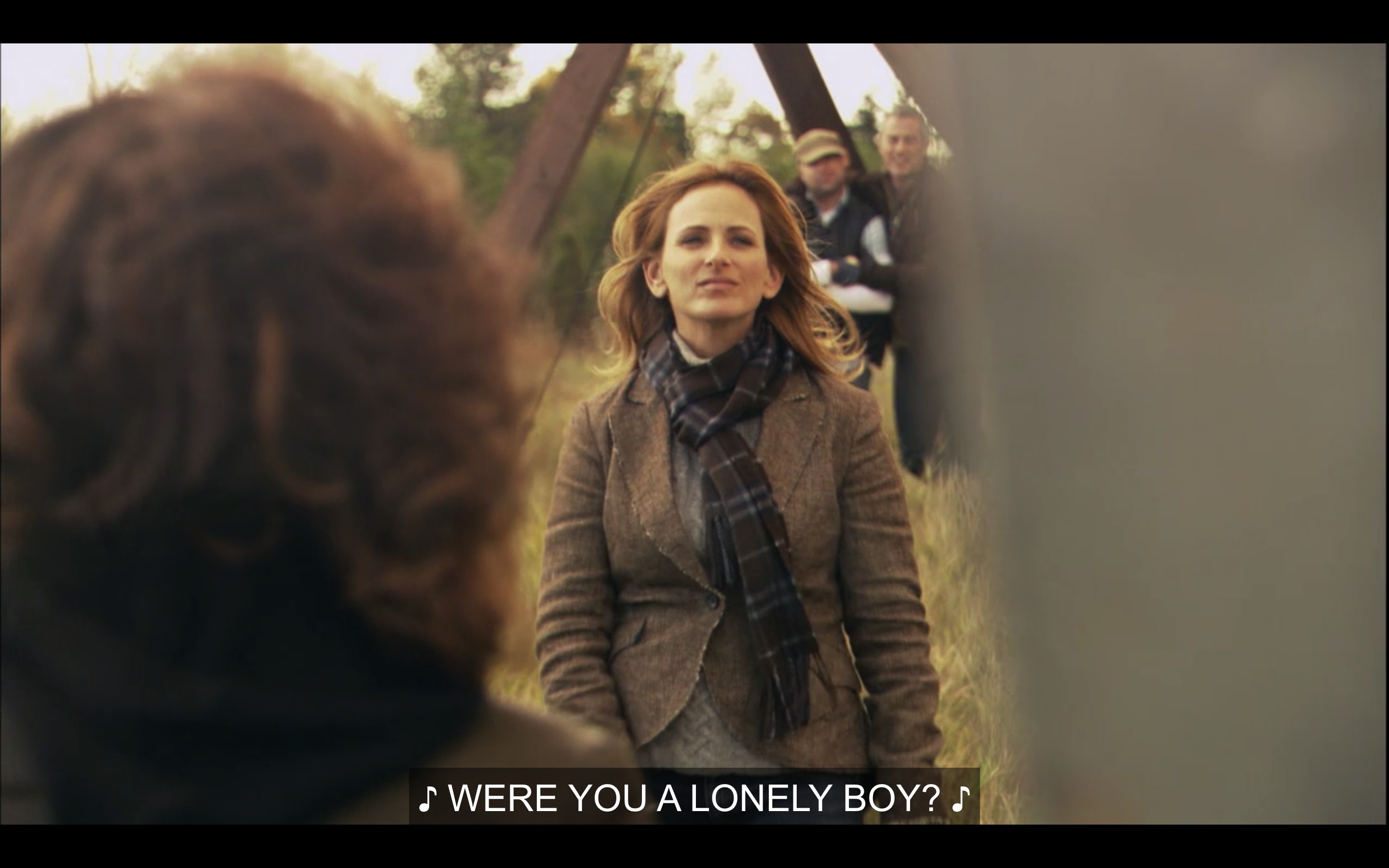 Jodi walks towards Bette (whose back is to the camera). Jodi is wearing a brown tweed blazer and a plaid scarf. They are outside. Music plays, singing, "Were you a lonely boy?"
