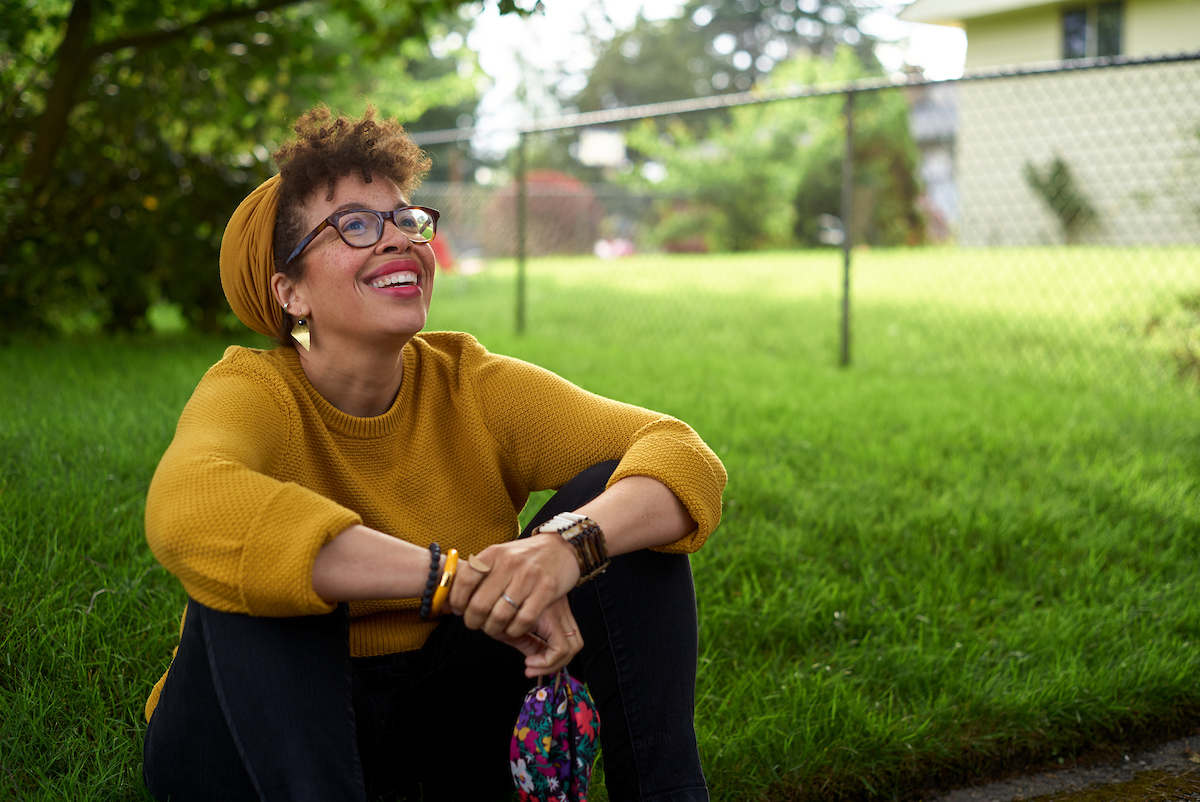 Kirsten Harris-Talley, who is running for public office in Seattle, sits on green grass in a yellow sweater and matching beanie, while she looks up into the sunlight.