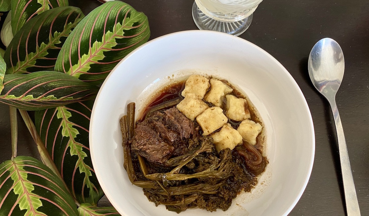 A bowl of braised beef, broccolini, and ricotta gnocchi sits on a black surface, and the leaves of a prayer plant snuggle up against it. A spoon sits next to the bowl and a cut crystal glass peeks into the edge of the frame. It's time to eat.