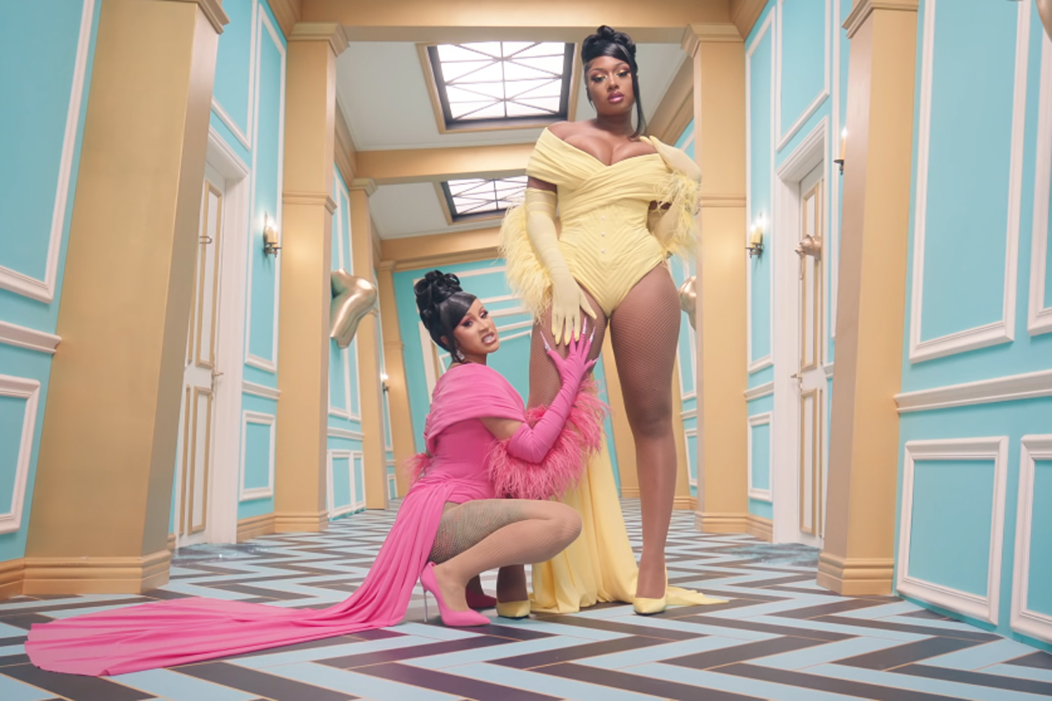 In the hallway of a mansion, Cardi B wears a pink bodysuit with a train, and kneels below Megan The Stallion, who stands prominently in the center, in a matching yellow bodysuit, looking like a boss. They both have gloved hands that meet on Meg's thigh, and are taunting the camera with sexily menacing stares..