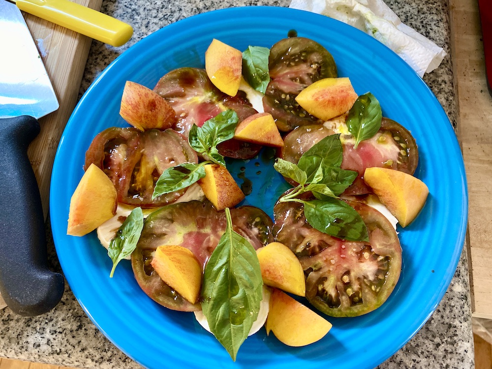 a bright, light blue plate with a circle of red tomatoes, orange nectarines, green basil leaves and peeks of white mozzarella underneath