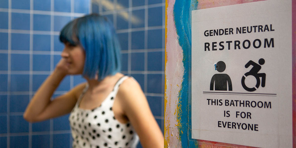 A nonbinary femme person with blue short hair stands in the background. In the foreground is a sign that reads, 