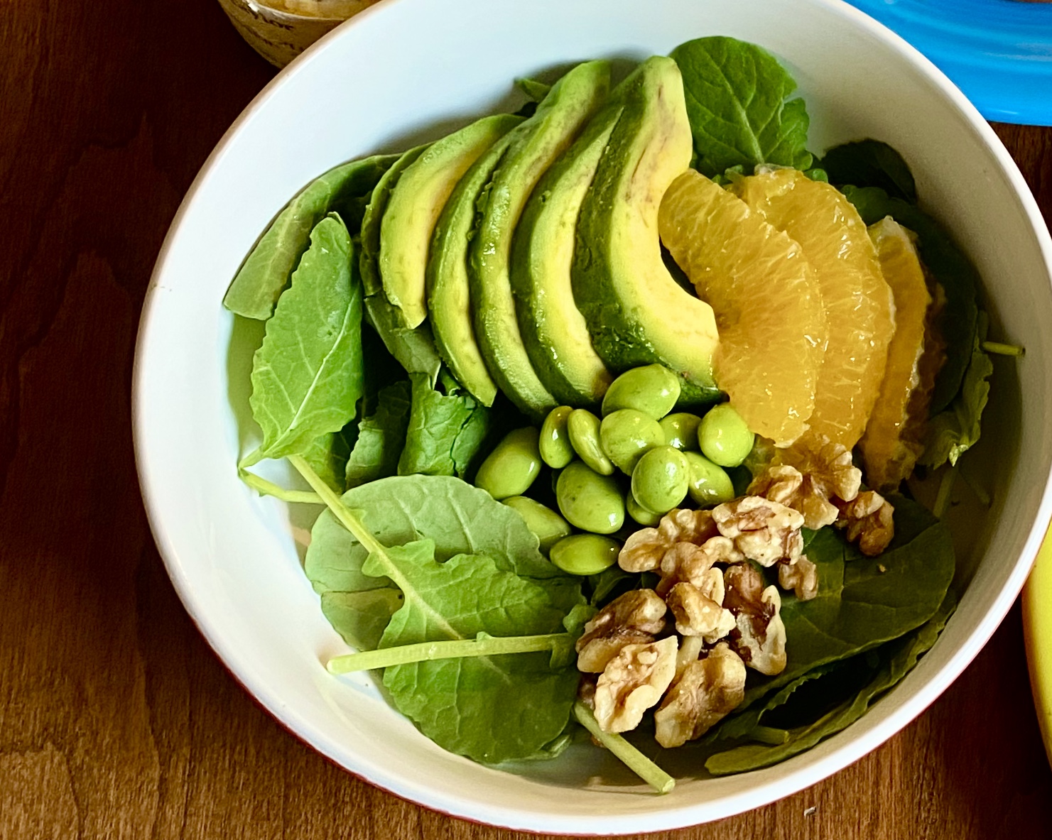 a shallow wide bowl with a bed of kale and fanned slices of avocado on top, slices of oranges nestled against those, a small pile of green edamame, and a small pile of walnuts