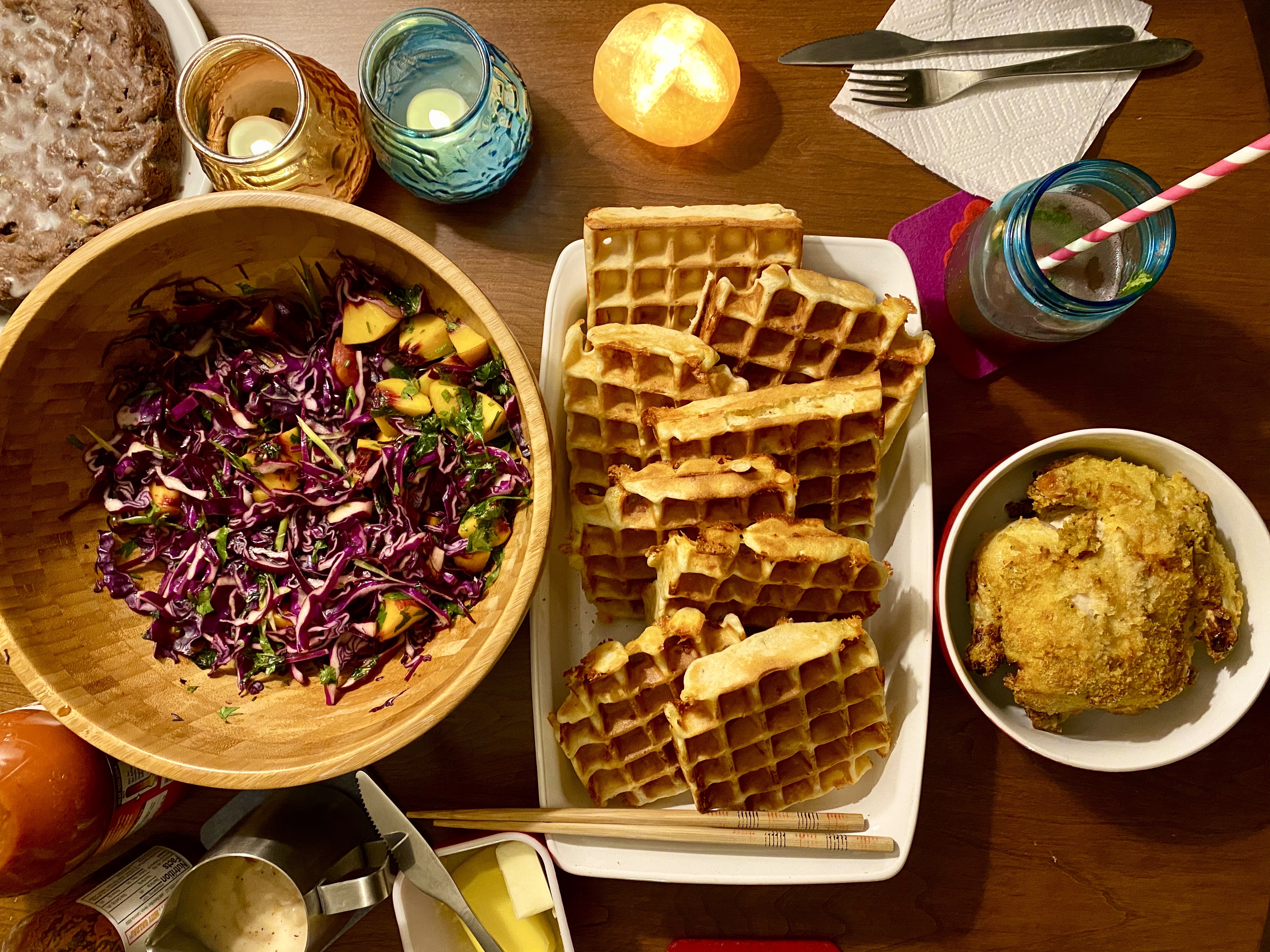 an overhead view of a table setting with a large bowl of purple cabbage salad, a tray of waffles, a pitcher of gravy, a small bowl with a little round fried hen nestled inside