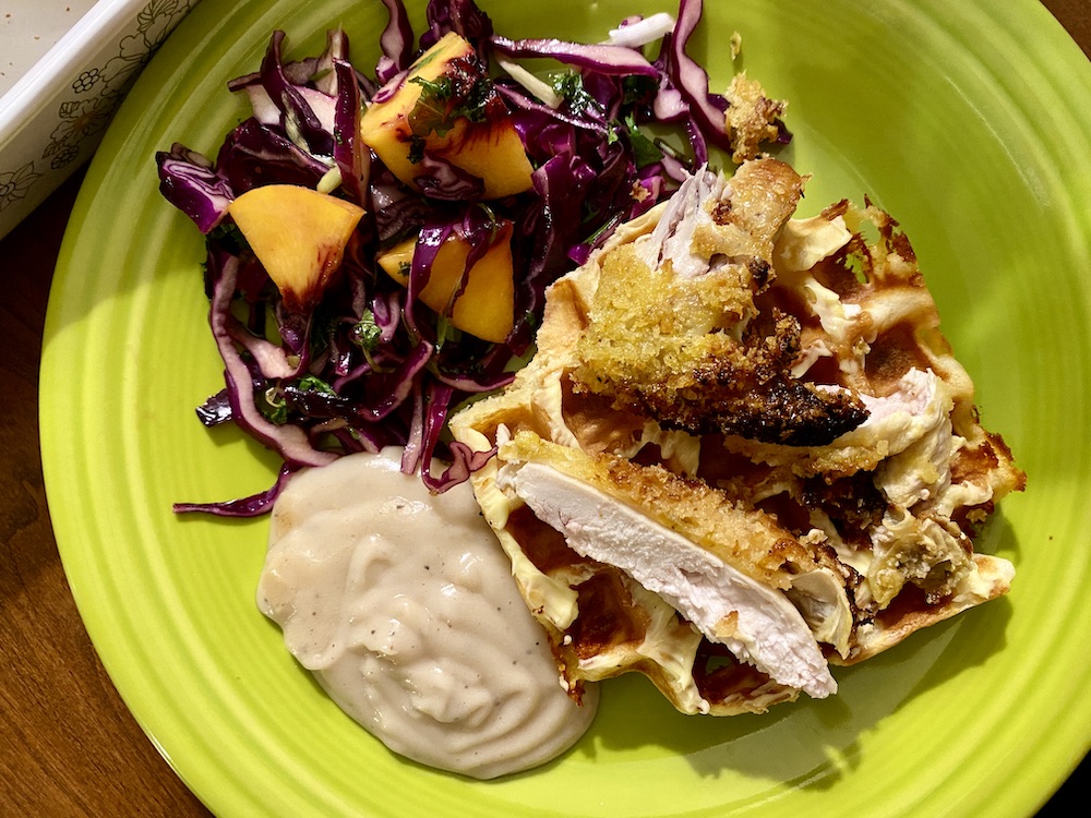 a bright green plate with a thick pool of gravy, a pile of purple cabbage salad w peach chunks, and a waffle and pieces of fried hen on top