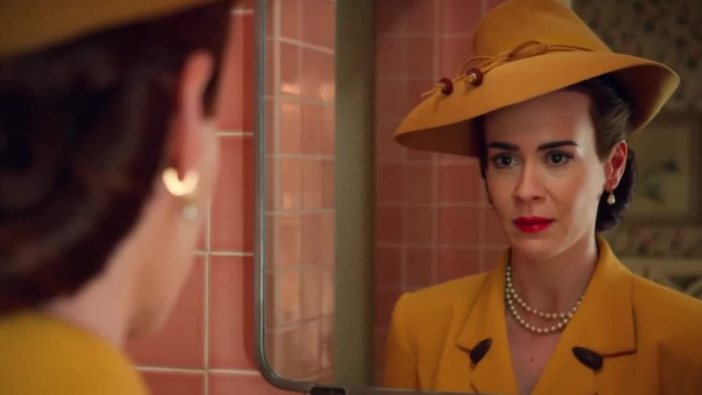 Sarah Paulson as Nurse Ratched in Ratched (showrunner Ryan Murphy, 2020). She is in a mustard yellow hat and dress styled like the 1940s and looks at herself in the mirror in a pink bathroom.