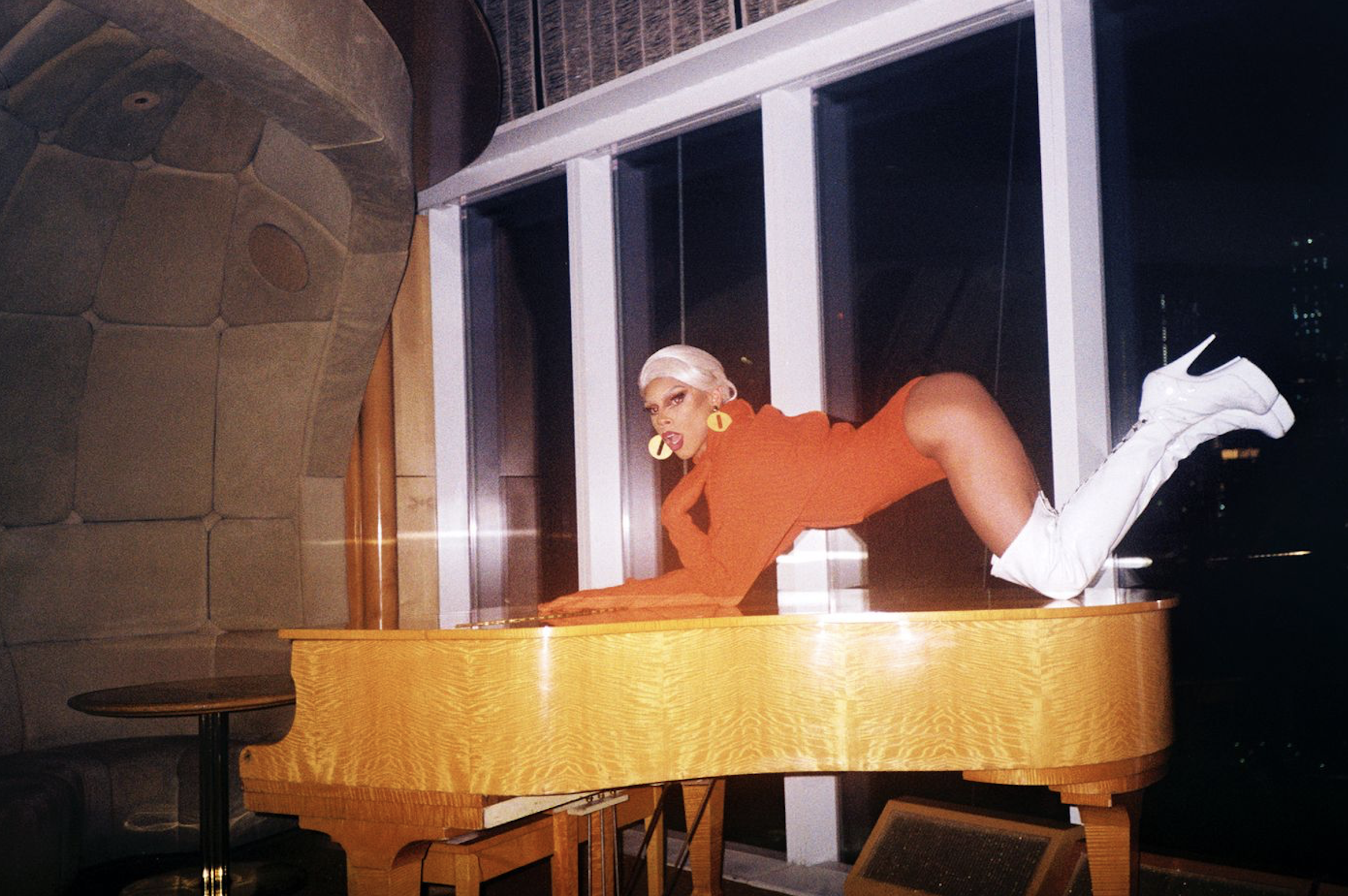 A Black trans woman with a white wig and an orange body suit, with thigh high white boots, crawls across a wooden piano.