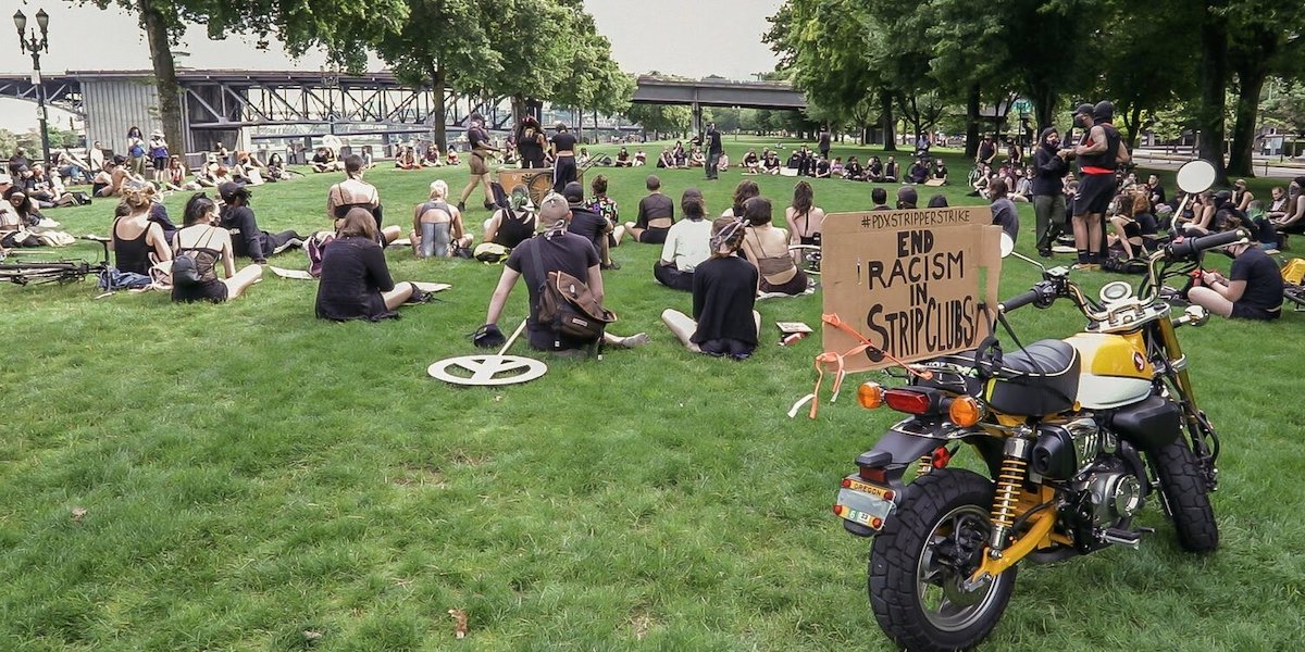 a group of people gathered on a lawn in portland, wearing masks and watching a few speakers in the middle. there is a sign taped to a motorcycle in the foreground that says "#pdxstripperstrike End Racism In Strip Clubs"