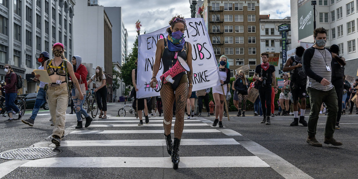 a group of people protesting in the street and soberly walking across a crosswalk. the person in the center wears black boots, black fishenets, a black body suit, a lilac scarf over their face and is carrying a hot pink and black cheetah megaphone across their body. they look downcast. behind them a protest sign half blocked by the people walking says 