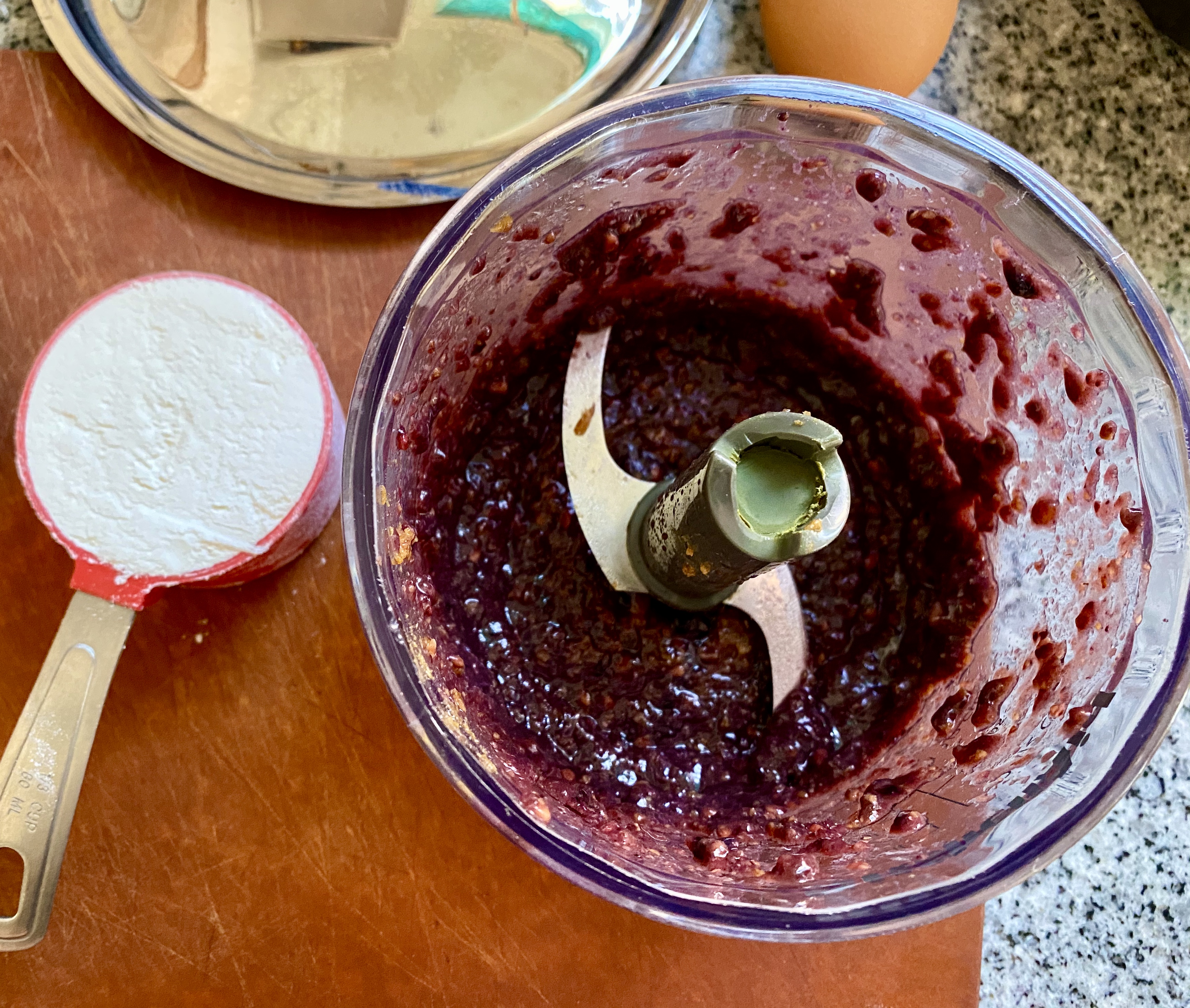 the bottom half of a mini food processor, the S-shaped blade, with blended blackberries and miso as a paste. a little 1/3 cup of flour sites beside the food processor.