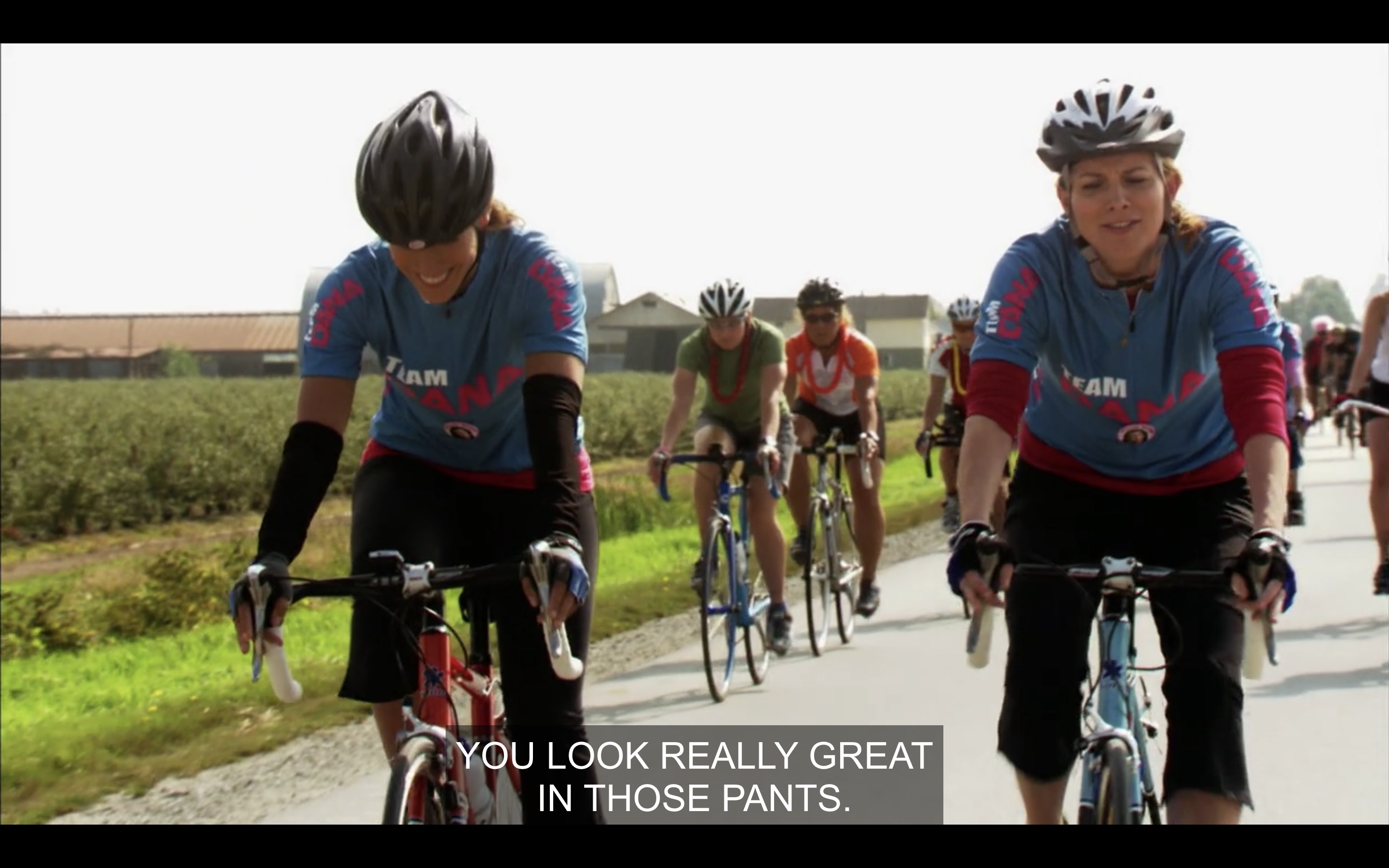 Bette and Tina biking, other bikers behind them, Pacific Northwest. Bette smiling, just told Tina her ass looks great in those pants