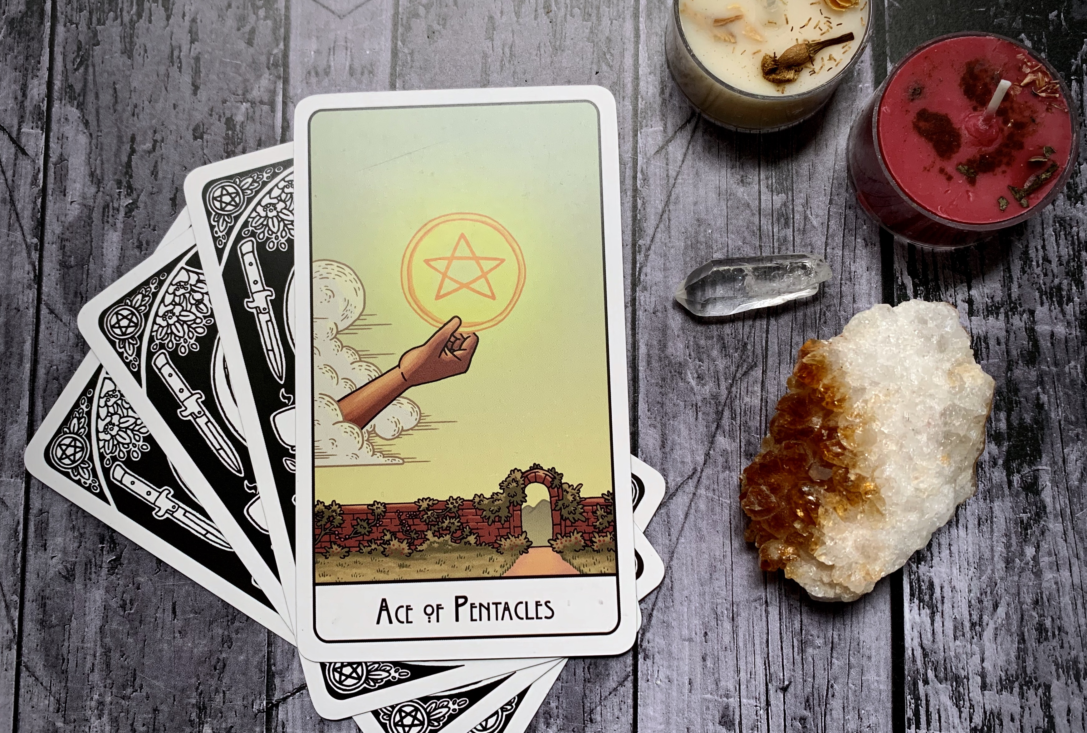 The Ace of Pentacles card sits on top of three other face-down cards. The Ace depicts a disembodied hand holding a glowing pentacle against a clear sky, below is a stone wall with a gate in it.