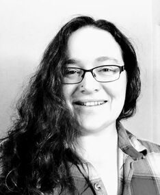 Black and white photo of G.B. Lindsey, who has long hair draped over one shoulder, glasses and a checked button-down shirt on. She is a contributor to Best Lesbian Erotica Vol 5