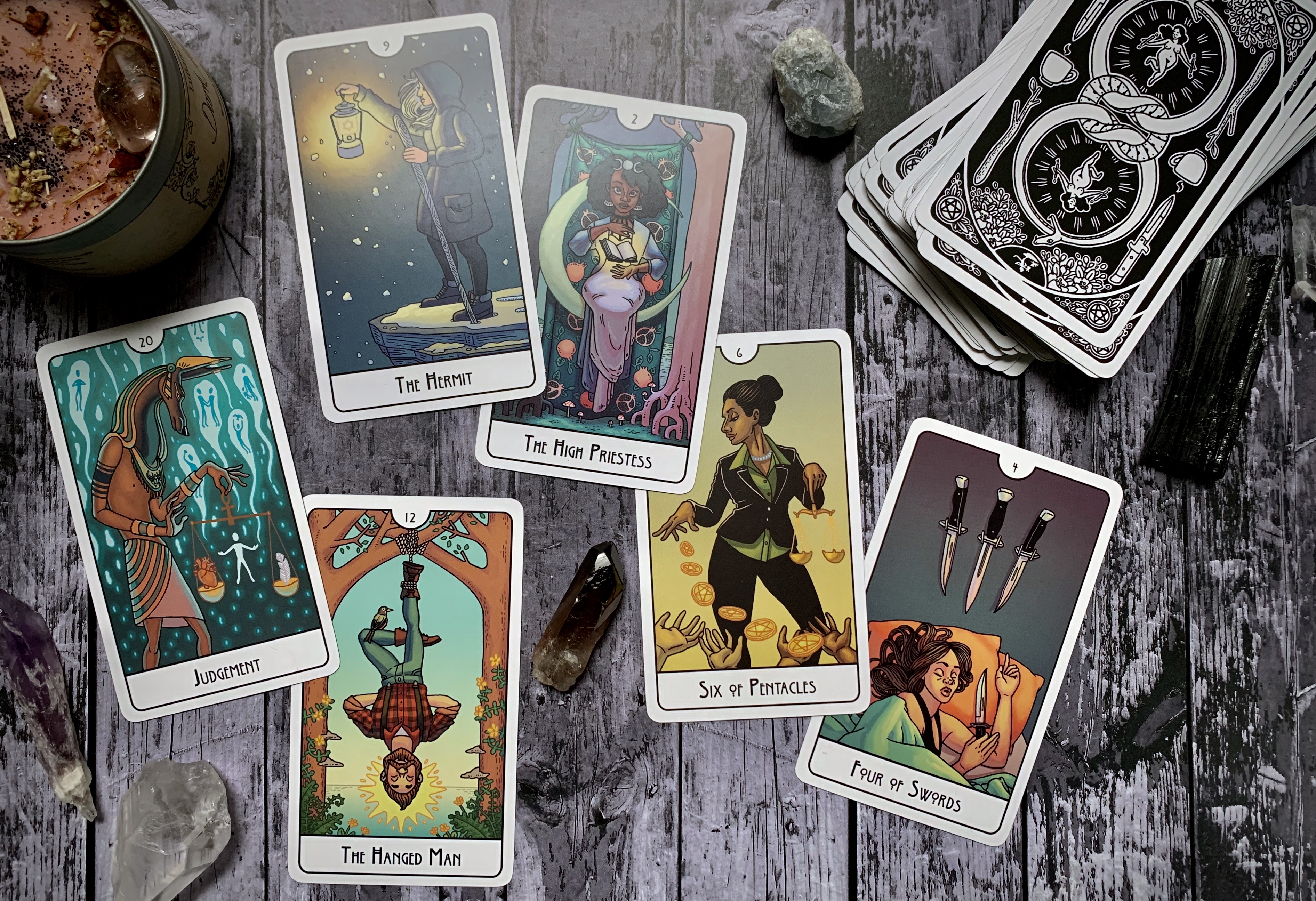There are six cards laid out in this photograph. Judgement depicts what appears to be the Egyptian god Anubis, holding a scale. On one end of the scale a human heart, on the other, a feather. In the background are a number of souls ascending against a teal backdrop. The Hermit card depicts a lone femme person with long silver or gray hair, in contemporary winter clothing, standing on the edge of a snowy cliff. They hold up a lantern and have a walking stick in hand. The Hanged Man card depicts a mas person in lumberjack-esque clothing, hanging upside down by their foot, from a tree. Their knee crosses, forming a triangle, and a bird rests atop their shin. The High Priestess card depicts a femme person, seated on a crescent moon, a glowing book floats between their hands. To the left is a white tree, and to the right is a dark brown tree. The Six of Pentacles card depicts a person in a business suit sprinkling gold coins to waiting hands below. Finally, the four of swords depicts a person asleep in bed, clutching one of the swords. The other three swords hang on the wall above them.