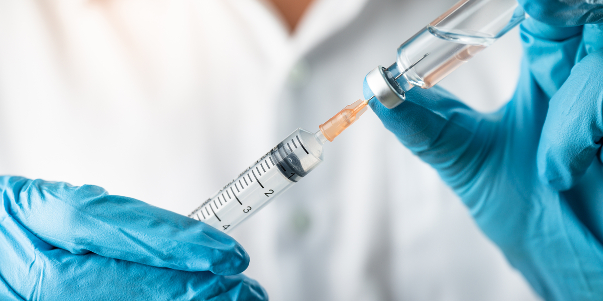 A pair of nitrile gloved hands fill an injection needle from a bottle.