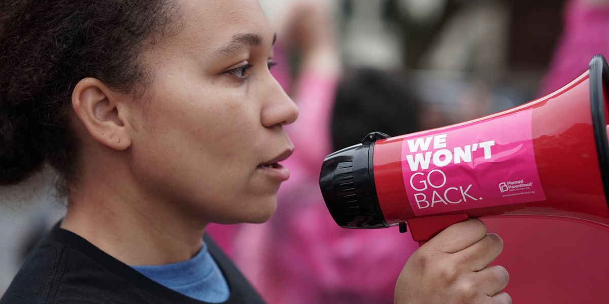 A close up photo portrait taken from the side of a woman with a serious expression on her face raising a megaphone to her mouth, with a pink Planned Parenthood sticker on it that reads WE WON'T GO BACK.