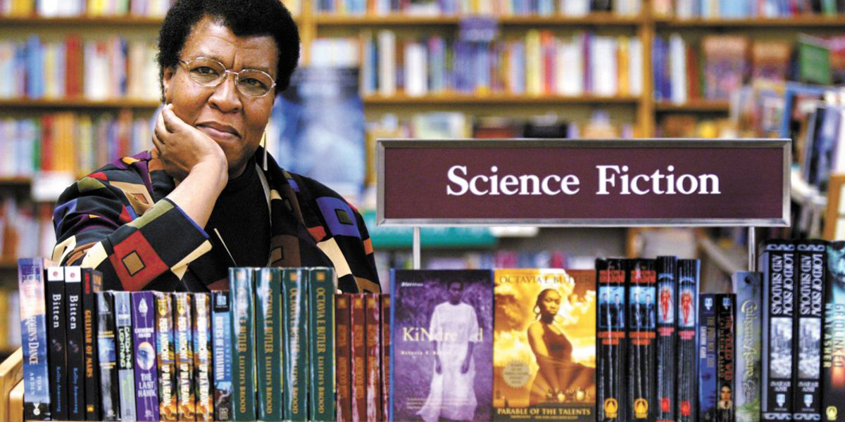 Octavia Butler in the 1990s stands in front of a collection of her books under the sign 
