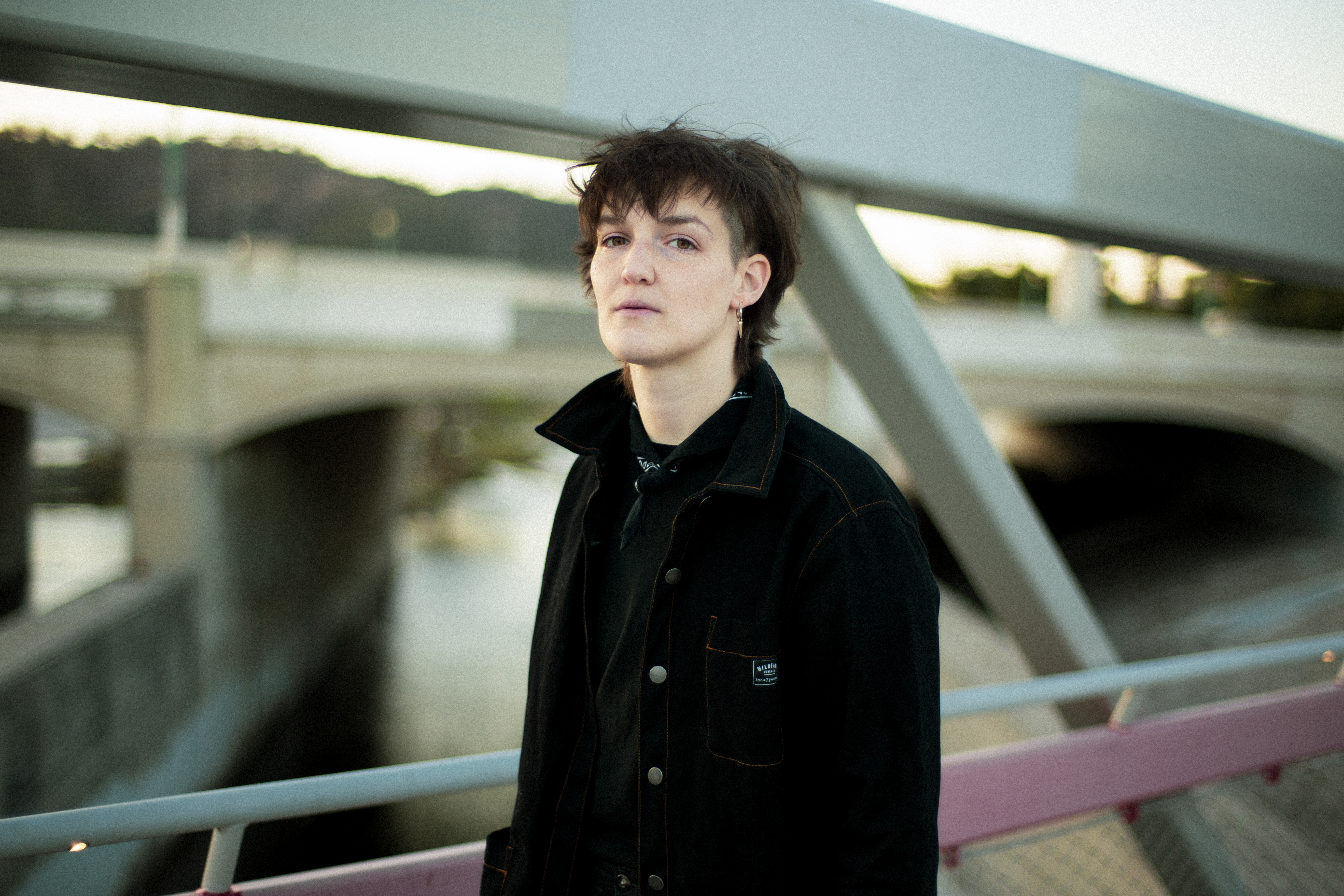 Artist Grace Semler Baldridge poses on a bridge. They are wearing a black coat and have a neutral expression.