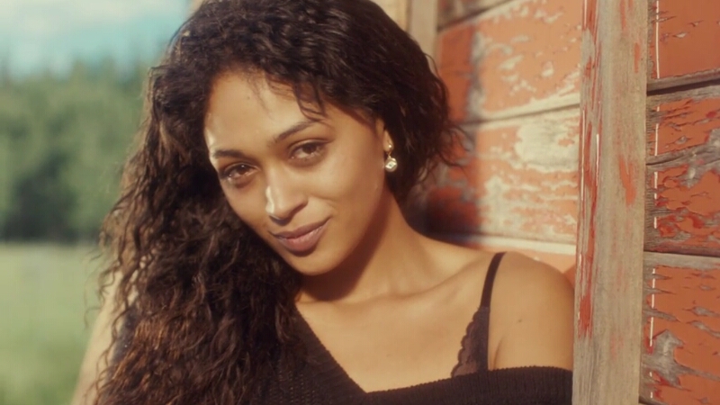 Cleo Clanton leans against a wood wall, one shoulder poking out of her shirt, head tilted, smiling softly and sweetly to camera as that 90s love glow washes over her.