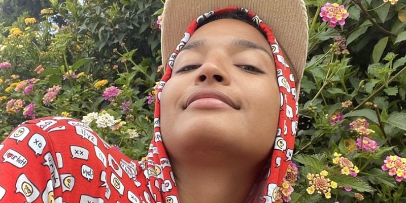 Indya Moore in a red hoodie sweatshirt against green plants and a Busch framing their face in the sunlight