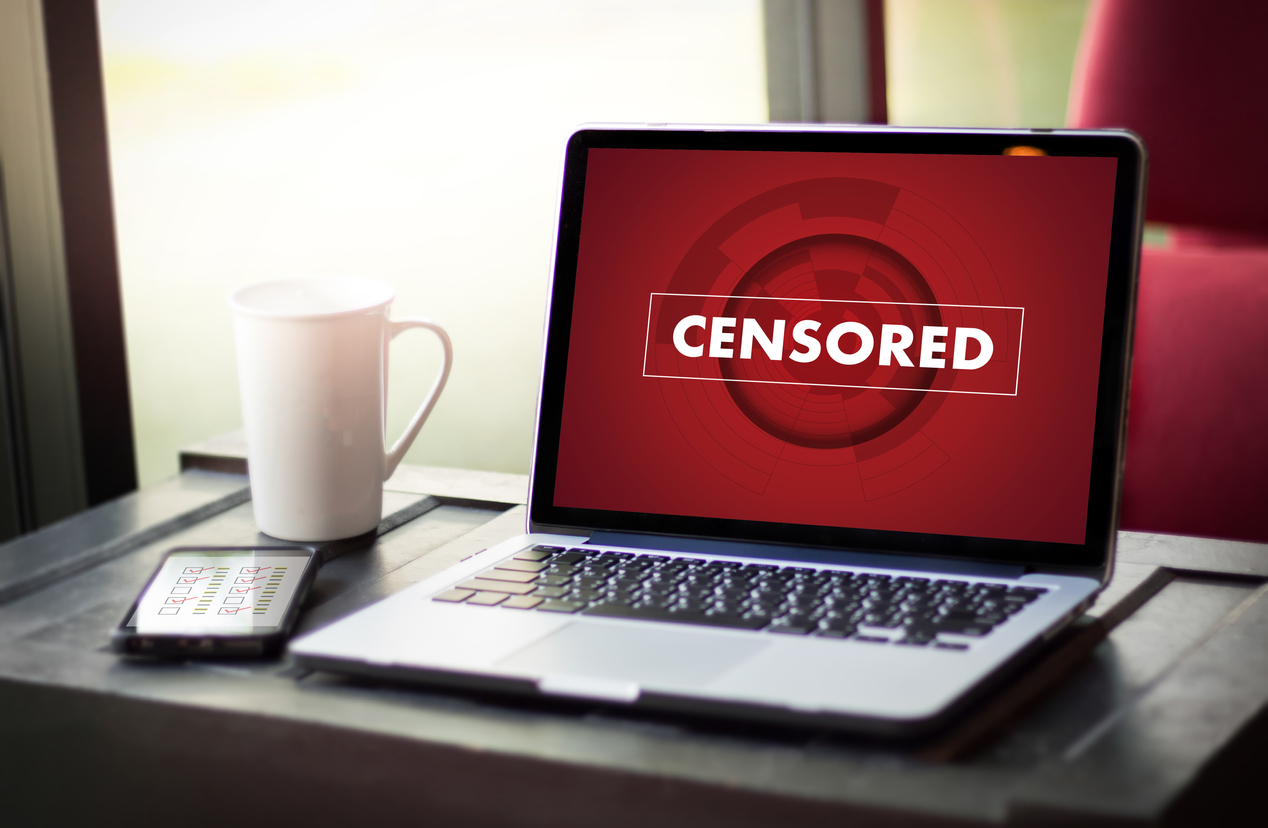 A photo of a laptop on a desktop with a coffee mug and smartphone; the laptop's screen displays a harsh red image that reads CENSORED