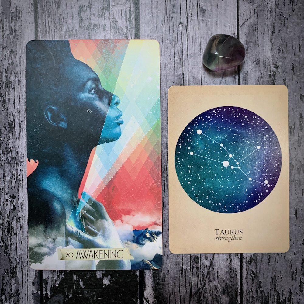 A photo of the tarot card Awakening, which features a Black woman's face in profile, the constellation card that read Taurus: Strengthen, and a grey crystal
