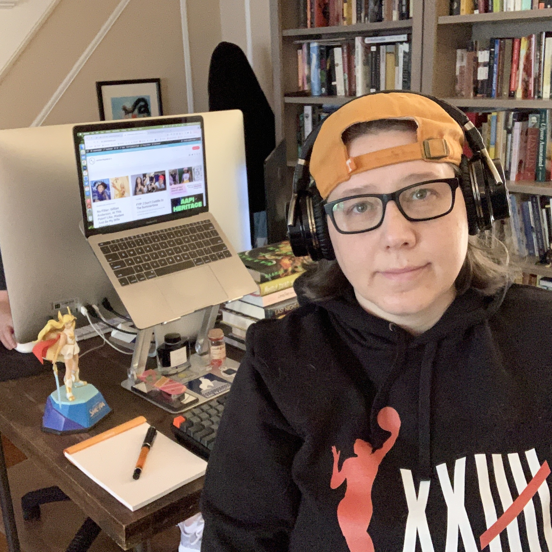 Heather Hogan sits at her desk, laptop open, with a small smile, a figure of she-ra in the backgound, and a glimpse of stacy's hand behind her. also, there is a notebook and pen on the desk and a stack of books. she has headphones, an orange hat, and a WNBA sweatshirt on